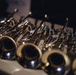 The image shows a collection of brass wind instruments, specifically trumpets, arranged closely together on a flat surface. The instruments display polished silver and gold finishes with characteristic bell openings facing the camera in the foreground. The shallow depth of field creates a bokeh effect, keeping only the nearest instruments in sharp focus.