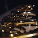The image shows multiple French horns arranged together, photographed in a dark, moody setting with dramatic lighting. The brass instruments display their characteristic coiled tubing and valves, with golden reflections and bokeh light effects creating an artistic composition. The shallow depth of field keeps the foreground instrument in partial focus while the background instruments blur into soft circles of light.