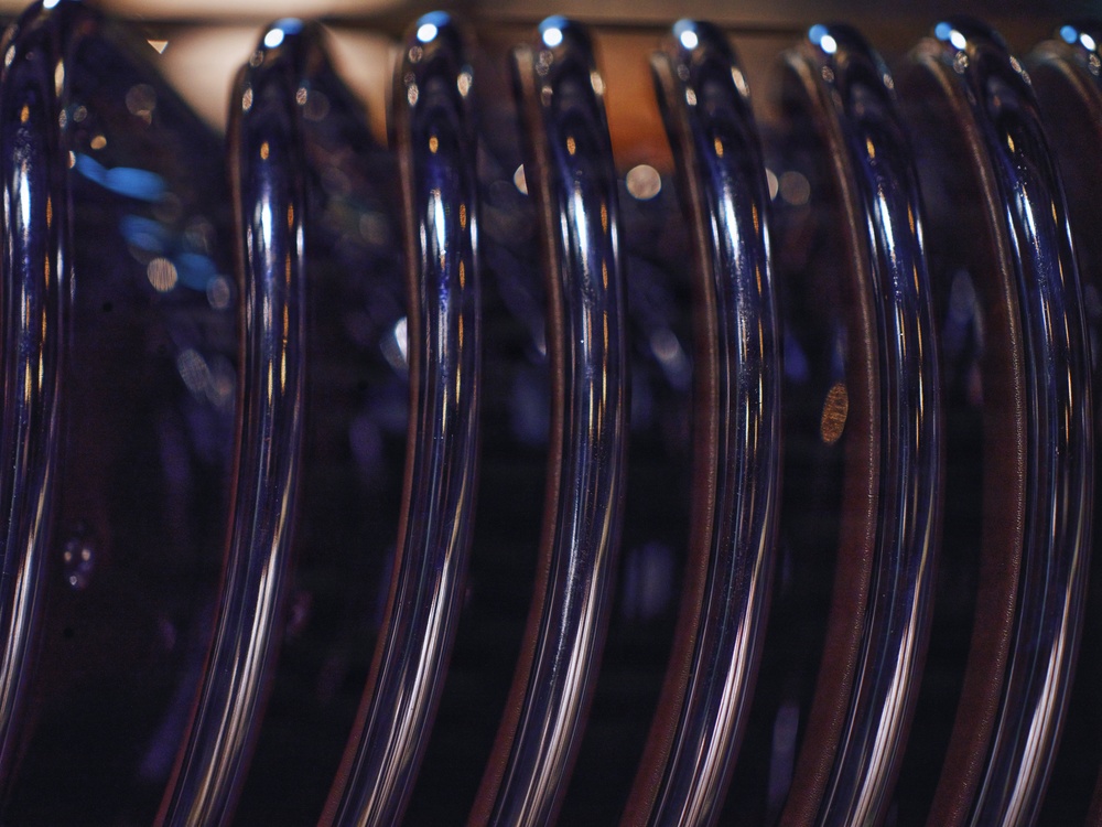 The image shows a close-up macro photograph of multiple clothes hangers lined up in a row. The hangers appear to be dark navy or black plastic with a glossy finish, reflecting ambient light as circular bokeh highlights. The shallow depth of field creates a blurred background while keeping the foreground hangers in sharp focus.
