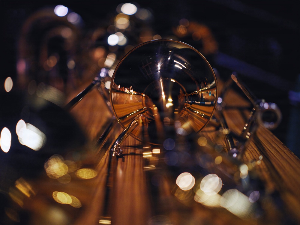 A close-up macro photograph of brass musical instruments, likely tubas or euphoniums, captured in a dimly lit environment. The central focus is on a polished bell of a brass instrument that reflects surrounding lights and instrument tubing in its curved surface. Bokeh light effects are visible throughout the background, creating an atmospheric, artistic composition.