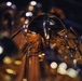 A close-up macro photograph of brass musical instruments, likely tubas or euphoniums, captured in a dimly lit environment. The central focus is on a polished bell of a brass instrument that reflects surrounding lights and instrument tubing in its curved surface. Bokeh light effects are visible throughout the background, creating an atmospheric, artistic composition.
