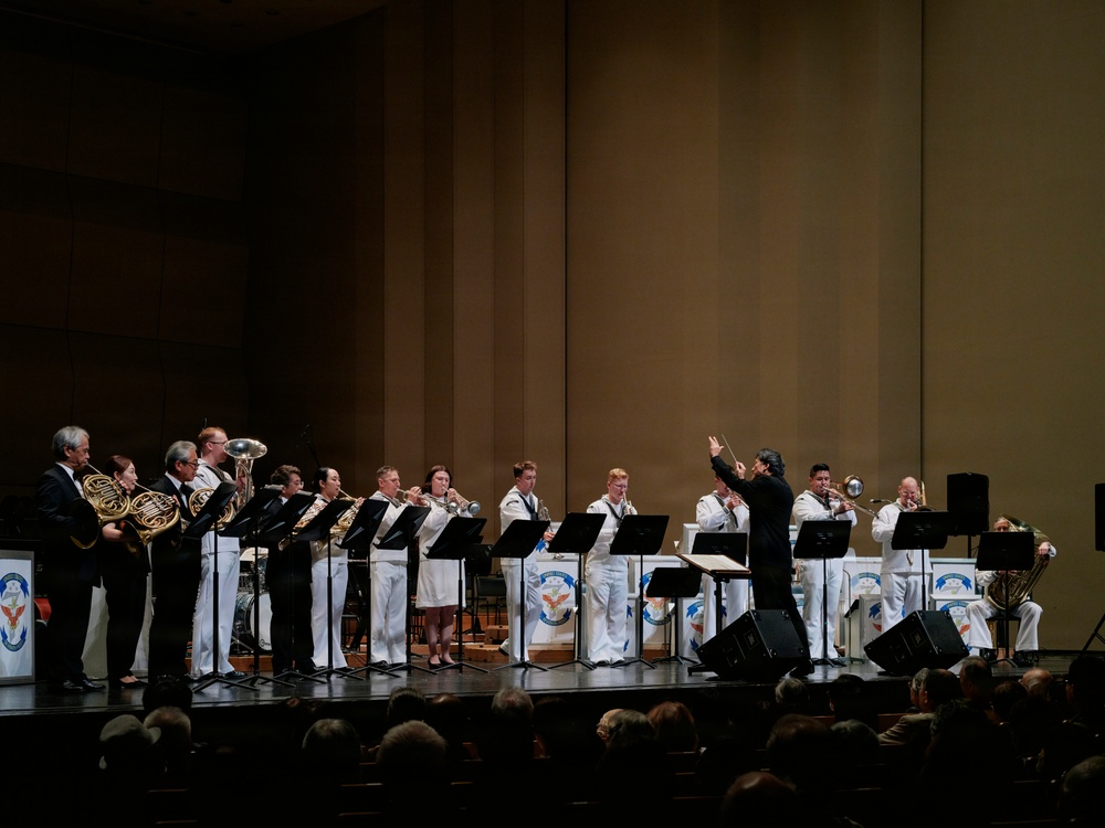 A military brass ensemble is performing on a concert hall stage, with musicians dressed in white naval uniforms and black formal attire. A conductor stands center-right, baton raised, leading the group. An audience is visible in the foreground watching the performance.