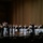 C7F Band Participates in American Sound in Japan A military brass ensemble is performing on a concert hall stage, with musicians dressed in white naval uniforms and black formal attire. A conductor stands center-right, baton raised, leading the group. An audience is visible in the foreground watching the performance.