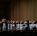 A military brass ensemble is performing on a concert hall stage, with musicians dressed in white naval uniforms and black formal attire. A conductor stands center-right, baton raised, leading the group. An audience is visible in the foreground watching the performance.