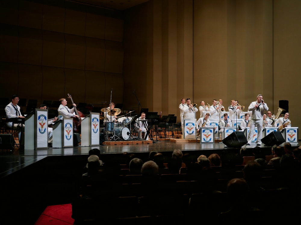A military band, likely the U.S. Navy Band, performs on a concert hall stage before a seated audience. The musicians are dressed in white naval dress uniforms and are arranged in a big band jazz ensemble configuration with brass, woodwind, rhythm section, and a featured soloist at the front. Music stands bear a logo featuring a bird emblem with red, white, and blue coloring.