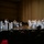C7F Band Participates in American Sound in Japan A military band, likely the U.S. Navy Band, performs on a concert hall stage before a seated audience. The musicians are dressed in white naval dress uniforms and are arranged in a big band jazz ensemble configuration with brass, woodwind, rhythm section, and a featured soloist at the front. Music stands bear a logo featuring a bird emblem with red, white, and blue coloring.