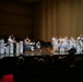 A military band, likely the U.S. Navy Band, performs on a concert hall stage before a seated audience. The musicians are dressed in white naval dress uniforms and are arranged in a big band jazz ensemble configuration with brass, woodwind, rhythm section, and a featured soloist at the front. Music stands bear a logo featuring a bird emblem with red, white, and blue coloring.