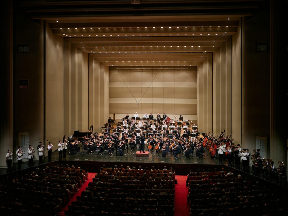 A large symphony orchestra is performing on a concert hall stage, with a conductor visible at the center podium. The venue appears to be a modern, well-appointed concert hall with warm beige acoustic paneling and rows of recessed ceiling lights. The audience is seated in red-carpeted seating areas, though attendance appears sparse with many empty seats.