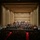 C7F Band Participates in American Sound in Japan A large symphony orchestra is performing on a concert hall stage, with a conductor visible at the center podium. The venue appears to be a modern, well-appointed concert hall with warm beige acoustic paneling and rows of recessed ceiling lights. The audience is seated in red-carpeted seating areas, though attendance appears sparse with many empty seats.