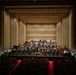 A large symphony orchestra is performing on a concert hall stage, with a conductor visible at the center podium. The venue appears to be a modern, well-appointed concert hall with warm beige acoustic paneling and rows of recessed ceiling lights. The audience is seated in red-carpeted seating areas, though attendance appears sparse with many empty seats.