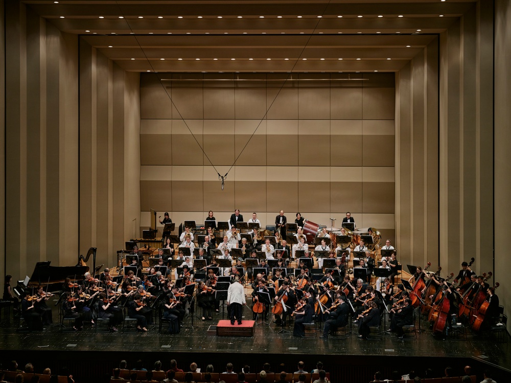 A full symphony orchestra is performing on a concert hall stage, led by a conductor standing on a red podium with their back to the camera. The musicians are arranged in the traditional orchestral semicircle formation across multiple tiers, dressed in formal black attire with the conductor in white. The modern concert hall features warm beige acoustic panels, recessed lighting, and a suspended microphone above the stage.