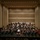 C7F Band Participates in American Sound in Japan A full symphony orchestra is performing on a concert hall stage, led by a conductor standing on a red podium with their back to the camera. The musicians are arranged in the traditional orchestral semicircle formation across multiple tiers, dressed in formal black attire with the conductor in white. The modern concert hall features warm beige acoustic panels, recessed lighting, and a suspended microphone above the stage.