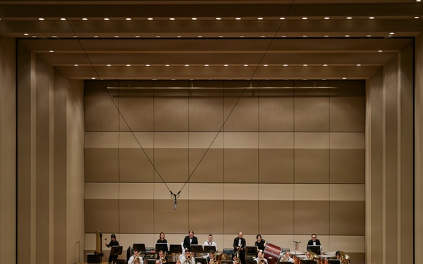 A full symphony orchestra is performing on a concert hall stage, led by a conductor standing on a red podium with their back to the camera. The musicians are arranged in the traditional orchestral semicircle formation across multiple tiers, dressed in formal black attire with the conductor in white. The modern concert hall features warm beige acoustic panels, recessed lighting, and a suspended microphone above the stage.