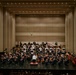 A full symphony orchestra is performing on a concert hall stage, led by a conductor standing on a red podium with their back to the camera. The musicians are arranged in the traditional orchestral semicircle formation across multiple tiers, dressed in formal black attire with the conductor in white. The modern concert hall features warm beige acoustic panels, recessed lighting, and a suspended microphone above the stage.