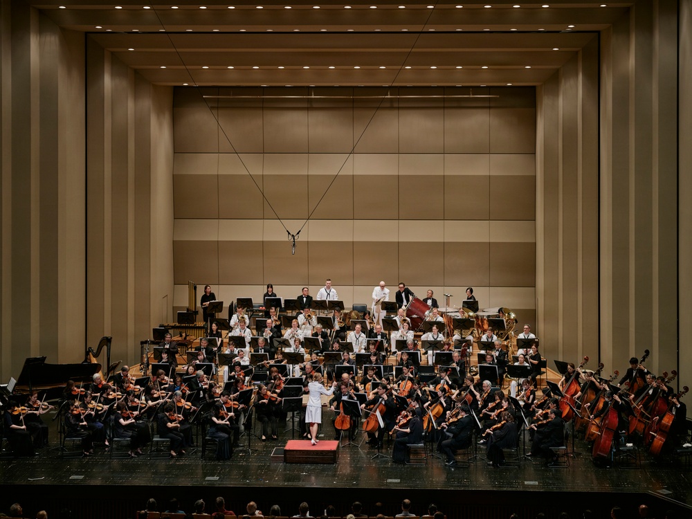 A full symphony orchestra is performing on a large concert hall stage, led by a conductor dressed in white standing on a red podium at center stage. The ensemble includes strings, brass, woodwinds, and percussion sections arranged in traditional orchestral formation. The modern concert hall features a neutral-toned acoustic wall backdrop with recessed ceiling lighting.
