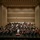 C7F Band Participates in American Sound in Japan A full symphony orchestra is performing on a large concert hall stage, led by a conductor dressed in white standing on a red podium at center stage. The ensemble includes strings, brass, woodwinds, and percussion sections arranged in traditional orchestral formation. The modern concert hall features a neutral-toned acoustic wall backdrop with recessed ceiling lighting.