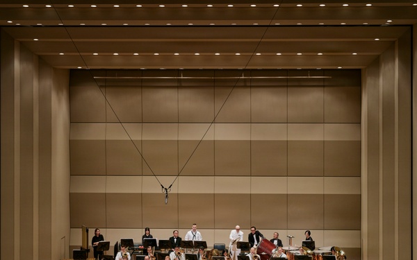 A full symphony orchestra is performing on a large concert hall stage, led by a conductor dressed in white standing on a red podium at center stage. The ensemble includes strings, brass, woodwinds, and percussion sections arranged in traditional orchestral formation. The modern concert hall features a neutral-toned acoustic wall backdrop with recessed ceiling lighting.