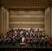 A full symphony orchestra is performing on a large concert hall stage, led by a conductor dressed in white standing on a red podium at center stage. The ensemble includes strings, brass, woodwinds, and percussion sections arranged in traditional orchestral formation. The modern concert hall features a neutral-toned acoustic wall backdrop with recessed ceiling lighting.