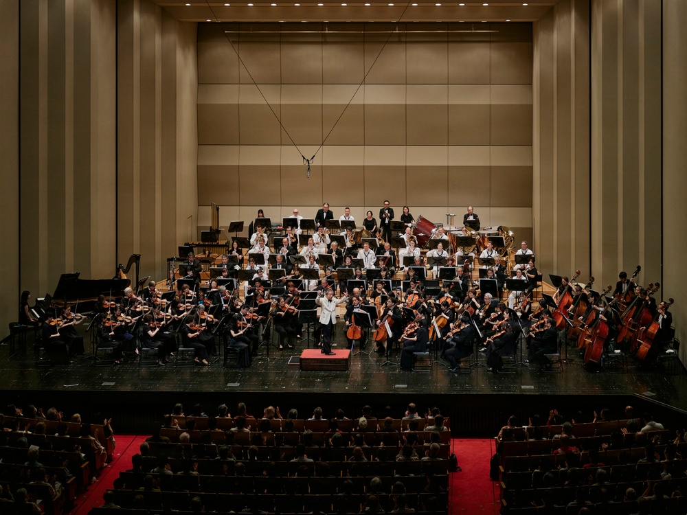 A full symphony orchestra is performing on a concert hall stage, with a conductor in a white jacket standing at the podium center stage. The ensemble includes string, woodwind, brass, and percussion sections arranged in traditional orchestral formation on a tiered stage. An audience is seated in the concert hall, and the venue features modern acoustic wall paneling.
