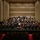 C7F Band Participates in American Sound in Japan A full symphony orchestra is performing on a concert hall stage, with a conductor in a white jacket standing at the podium center stage. The ensemble includes string, woodwind, brass, and percussion sections arranged in traditional orchestral formation on a tiered stage. An audience is seated in the concert hall, and the venue features modern acoustic wall paneling.