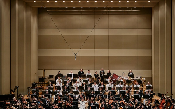 A full symphony orchestra is performing on a concert hall stage, with a conductor in a white jacket standing at the podium center stage. The ensemble includes string, woodwind, brass, and percussion sections arranged in traditional orchestral formation on a tiered stage. An audience is seated in the concert hall, and the venue features modern acoustic wall paneling.