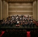 A full symphony orchestra is performing on a concert hall stage, with a conductor in a white jacket standing at the podium center stage. The ensemble includes string, woodwind, brass, and percussion sections arranged in traditional orchestral formation on a tiered stage. An audience is seated in the concert hall, and the venue features modern acoustic wall paneling.