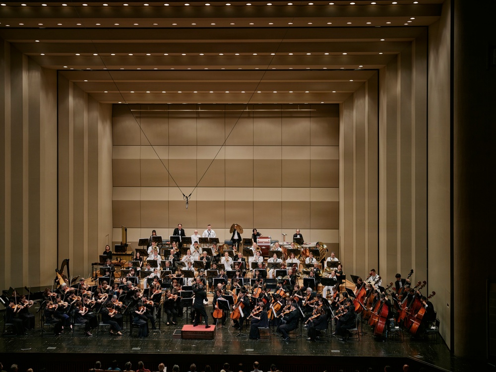 A full symphony orchestra is performing on a large concert hall stage, with a conductor standing on a podium at the center front. The musicians are arranged in the traditional orchestral layout across multiple tiered levels, dressed in formal black attire with some wind/brass players in white. The venue features a modern, acoustically designed hall with warm beige paneling, recessed lighting, and suspended microphones.