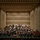 C7F Band Participates in American Sound in Japan A full symphony orchestra is performing on a large concert hall stage, with a conductor standing on a podium at the center front. The musicians are arranged in the traditional orchestral layout across multiple tiered levels, dressed in formal black attire with some wind/brass players in white. The venue features a modern, acoustically designed hall with warm beige paneling, recessed lighting, and suspended microphones.
