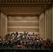 A full symphony orchestra is performing on a large concert hall stage, with a conductor standing on a podium at the center front. The musicians are arranged in the traditional orchestral layout across multiple tiered levels, dressed in formal black attire with some wind/brass players in white. The venue features a modern, acoustically designed hall with warm beige paneling, recessed lighting, and suspended microphones.