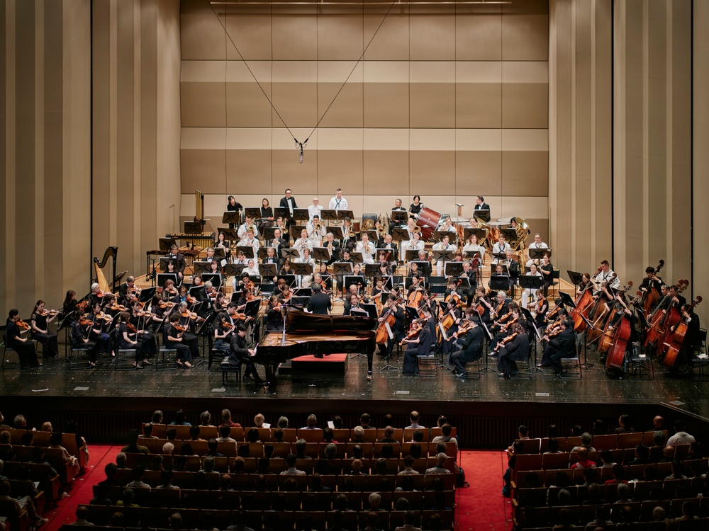 A full symphony orchestra is performing on a concert hall stage, with a conductor standing at the podium and a pianist seated at a grand piano, suggesting a piano concerto performance. The large ensemble includes strings, brass, woodwinds, and percussion sections arranged in traditional orchestral formation. The audience is seated in red-carpeted auditorium seating, watching the performance from the front rows.