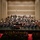C7F Band Participates in American Sound in Japan A full symphony orchestra is performing on a concert hall stage, with a conductor standing at the podium and a pianist seated at a grand piano, suggesting a piano concerto performance. The large ensemble includes strings, brass, woodwinds, and percussion sections arranged in traditional orchestral formation. The audience is seated in red-carpeted auditorium seating, watching the performance from the front rows.