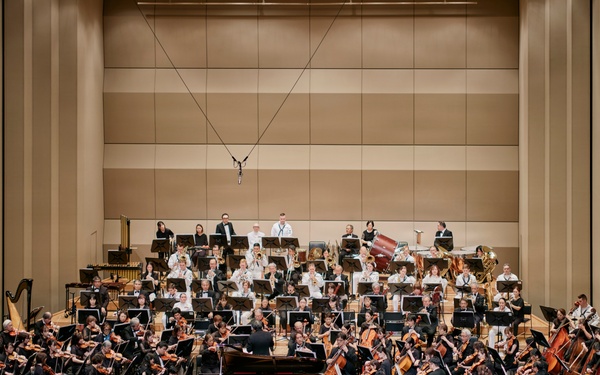 A full symphony orchestra is performing on a concert hall stage, with a conductor standing at the podium and a pianist seated at a grand piano, suggesting a piano concerto performance. The large ensemble includes strings, brass, woodwinds, and percussion sections arranged in traditional orchestral formation. The audience is seated in red-carpeted auditorium seating, watching the performance from the front rows.