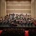 A full symphony orchestra is performing on a concert hall stage, with a conductor standing at the podium and a pianist seated at a grand piano, suggesting a piano concerto performance. The large ensemble includes strings, brass, woodwinds, and percussion sections arranged in traditional orchestral formation. The audience is seated in red-carpeted auditorium seating, watching the performance from the front rows.
