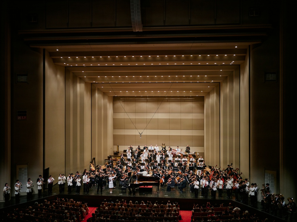 A large symphony orchestra is assembled on a concert hall stage, appearing to be taking a bow or concluding a performance. The stage features a grand piano as a centerpiece, with musicians arranged in traditional orchestral seating tiers. The venue is a modern concert hall with warm beige acoustic paneling and rows of red-seated audience chairs visible in the foreground.