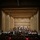C7F Band Participates in American Sound in Japan A large symphony orchestra is assembled on a concert hall stage, appearing to be taking a bow or concluding a performance. The stage features a grand piano as a centerpiece, with musicians arranged in traditional orchestral seating tiers. The venue is a modern concert hall with warm beige acoustic paneling and rows of red-seated audience chairs visible in the foreground.