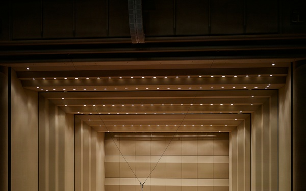 A large symphony orchestra is assembled on a concert hall stage, appearing to be taking a bow or concluding a performance. The stage features a grand piano as a centerpiece, with musicians arranged in traditional orchestral seating tiers. The venue is a modern concert hall with warm beige acoustic paneling and rows of red-seated audience chairs visible in the foreground.