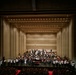 A large symphony orchestra is assembled on a concert hall stage, appearing to be taking a bow or concluding a performance. The stage features a grand piano as a centerpiece, with musicians arranged in traditional orchestral seating tiers. The venue is a modern concert hall with warm beige acoustic paneling and rows of red-seated audience chairs visible in the foreground.