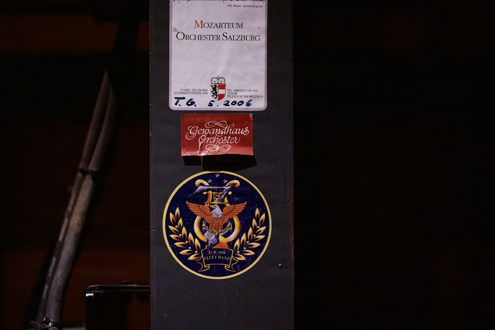 The image shows a dark surface, likely a music stand or equipment case, with three stickers/labels affixed to it. The labels represent different musical organizations: the Mozarteum Orchester Salzburg, the Gewandhaus Orchester, and the U.S. 7th Fleet Band. A handwritten date '5. 2006' is visible on the Mozarteum label.