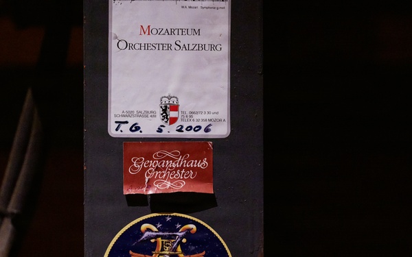 The image shows a dark surface, likely a music stand or equipment case, with three stickers/labels affixed to it. The labels represent different musical organizations: the Mozarteum Orchester Salzburg, the Gewandhaus Orchester, and the U.S. 7th Fleet Band. A handwritten date '5. 2006' is visible on the Mozarteum label.