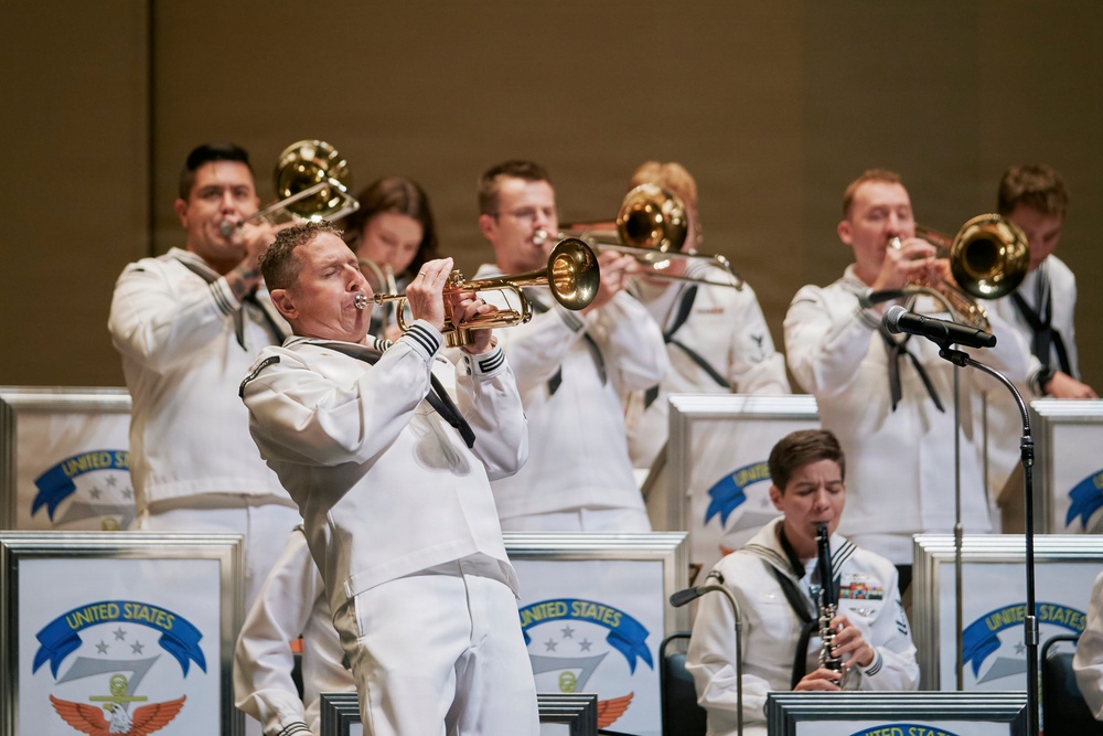 A United States Navy band is performing in formal white dress uniforms on a stage. Multiple musicians are playing brass instruments including trumpets and trombones, with at least one woodwind player visible in the foreground. Music stands bearing the 'United States' insignia with an eagle emblem are positioned in front of the performers.