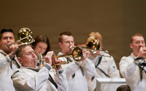 A United States Navy band is performing in formal white dress uniforms on a stage. Multiple musicians are playing brass instruments including trumpets and trombones, with at least one woodwind player visible in the foreground. Music stands bearing the 'United States' insignia with an eagle emblem are positioned in front of the performers.