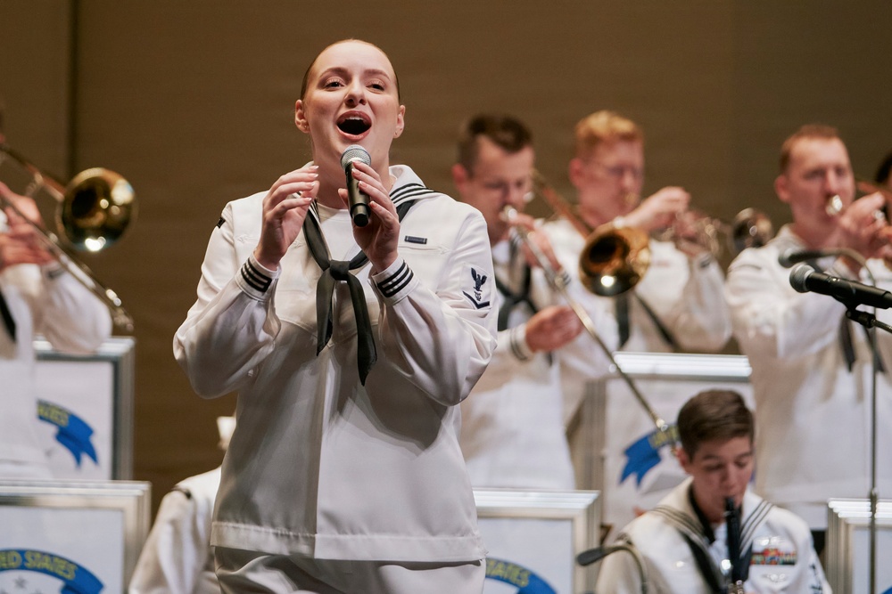 A female U.S. Navy sailor performs as a vocalist at a military band concert, holding a microphone and singing in the foreground. Behind her, multiple male sailors in matching white dress uniforms play brass instruments including trumpets and trombones. Music stands with partial 'United States' text are visible, indicating this is an official U.S. Navy Band performance.