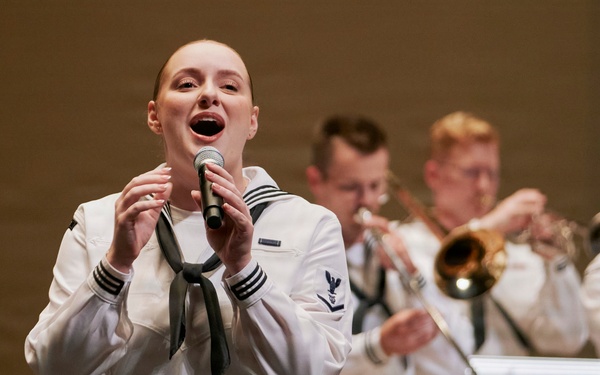 A female U.S. Navy sailor performs as a vocalist at a military band concert, holding a microphone and singing in the foreground. Behind her, multiple male sailors in matching white dress uniforms play brass instruments including trumpets and trombones. Music stands with partial 'United States' text are visible, indicating this is an official U.S. Navy Band performance.