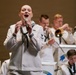 A female U.S. Navy sailor performs as a vocalist at a military band concert, holding a microphone and singing in the foreground. Behind her, multiple male sailors in matching white dress uniforms play brass instruments including trumpets and trombones. Music stands with partial 'United States' text are visible, indicating this is an official U.S. Navy Band performance.