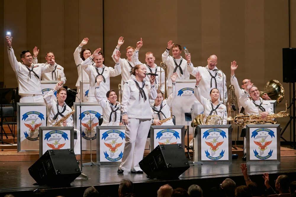 A United States Navy Seventh Fleet band performs on stage in a concert hall, with members dressed in traditional white Navy uniforms. A female vocalist stands at center stage singing into a microphone while band members behind her enthusiastically raise their hands. Music stands with the United States Seventh Fleet insignia are prominently displayed across the front of the stage.