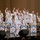 C7F Band Participates in American Sound in Japan A United States Navy Seventh Fleet band performs on stage in a concert hall, with members dressed in traditional white Navy uniforms. A female vocalist stands at center stage singing into a microphone while band members behind her enthusiastically raise their hands. Music stands with the United States Seventh Fleet insignia are prominently displayed across the front of the stage.