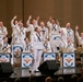 A United States Navy Seventh Fleet band performs on stage in a concert hall, with members dressed in traditional white Navy uniforms. A female vocalist stands at center stage singing into a microphone while band members behind her enthusiastically raise their hands. Music stands with the United States Seventh Fleet insignia are prominently displayed across the front of the stage.