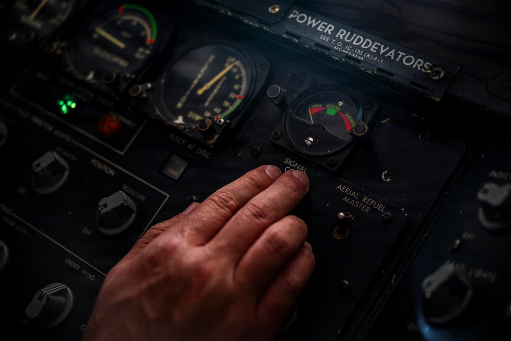 A close-up photograph of an aircraft cockpit control panel, showing a human hand operating controls near labeled switches and gauges. The panel includes markings for 'POWER RUDDEVATORS,' 'AERIAL REFUELING MASTER,' and 'SIGNAL' controls, consistent with a military tanker or transport aircraft. The reference to T.O. 1C-135(K)A-1 and BAC-M93-60-8A suggests this is a KC-135 Stratotanker cockpit.