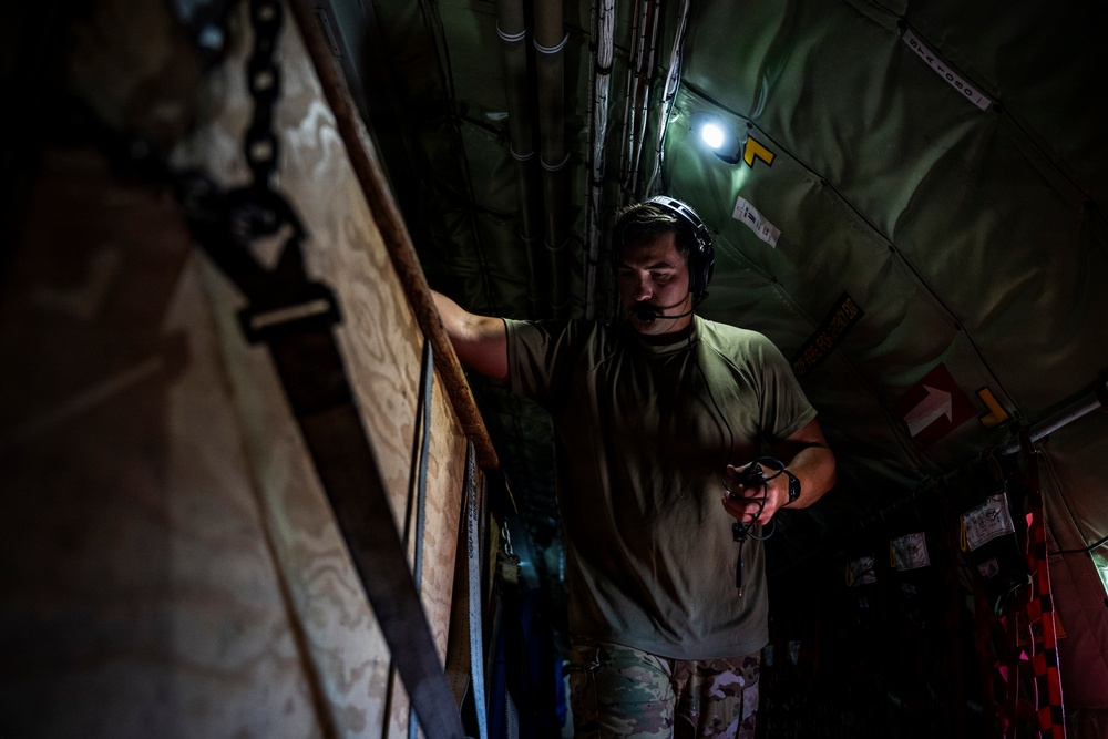 A military personnel member is standing inside what appears to be the cargo hold of a military aircraft. The individual is wearing a communication headset and olive drab t-shirt while examining something in their hands. The interior shows typical military aircraft cargo bay features including chains, straps, pipes, and warning labels.