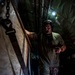 A military personnel member is standing inside what appears to be the cargo hold of a military aircraft. The individual is wearing a communication headset and olive drab t-shirt while examining something in their hands. The interior shows typical military aircraft cargo bay features including chains, straps, pipes, and warning labels.